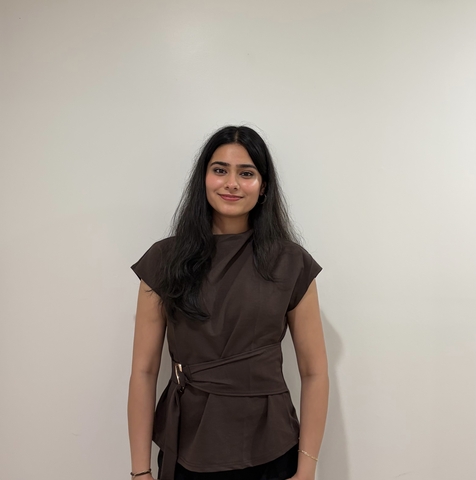 A woman with long black hair wearing a brown blouse standing against a white wall.