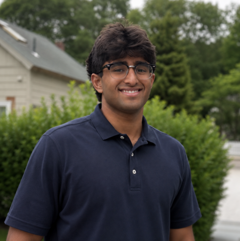 Man in glasses smiling in front of green bushes.