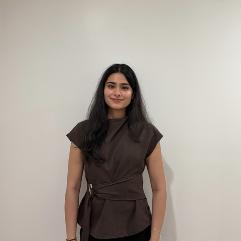 A woman with long black hair wearing a brown blouse standing against a white wall.